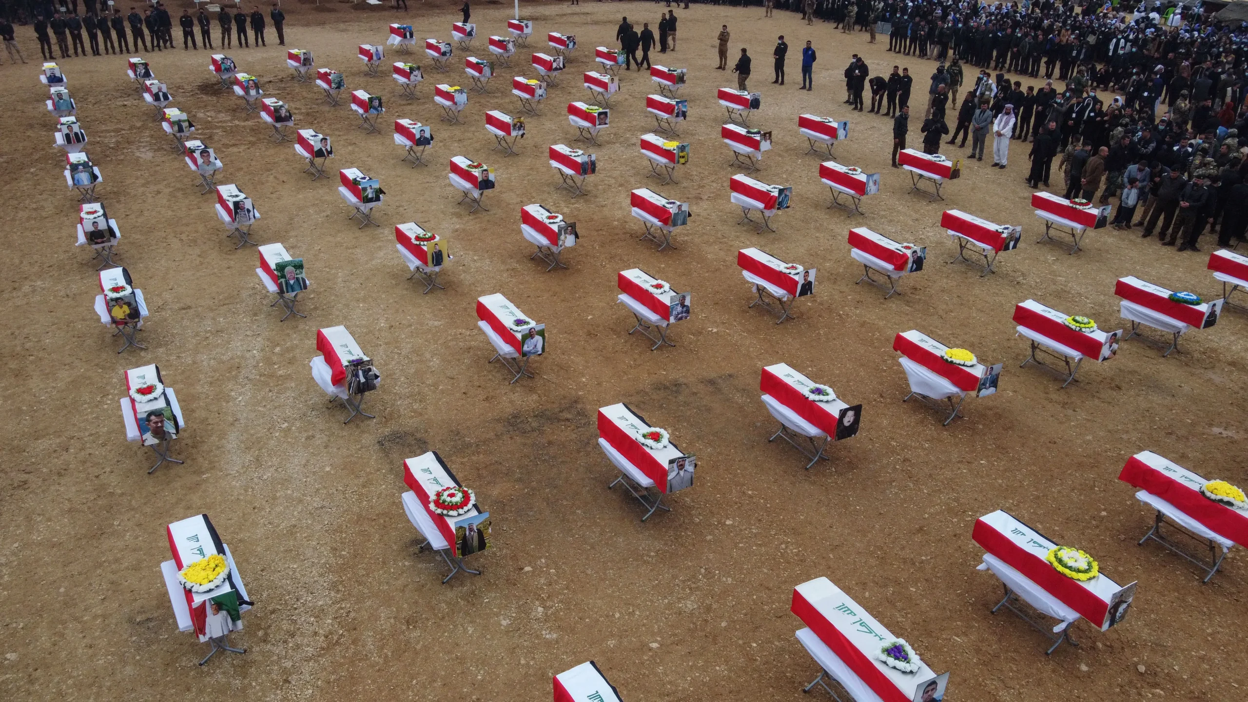 Rows of flag-draped coffins laid out on open ground at a funeral, with mourners gathered behind them